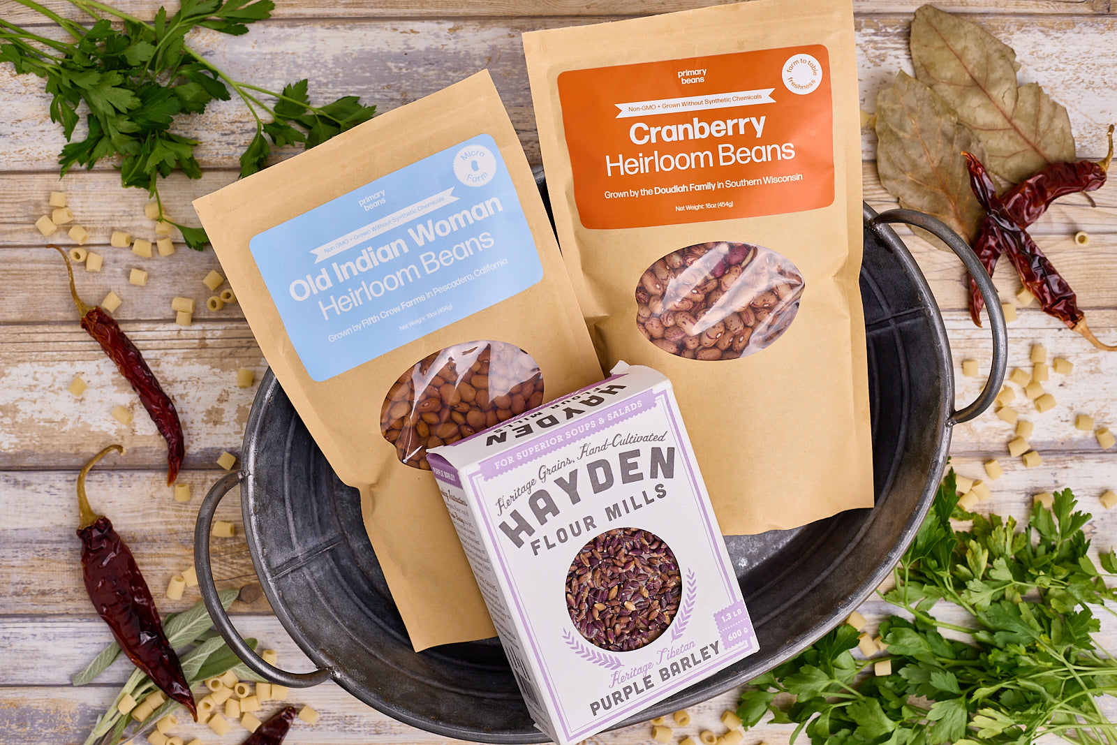 January Bean and Grain Club ingredients with two bags of heirloom beans and one box of purple barley grains inside a metal tin on a wooden counter surrounded by decorative herbs and spices