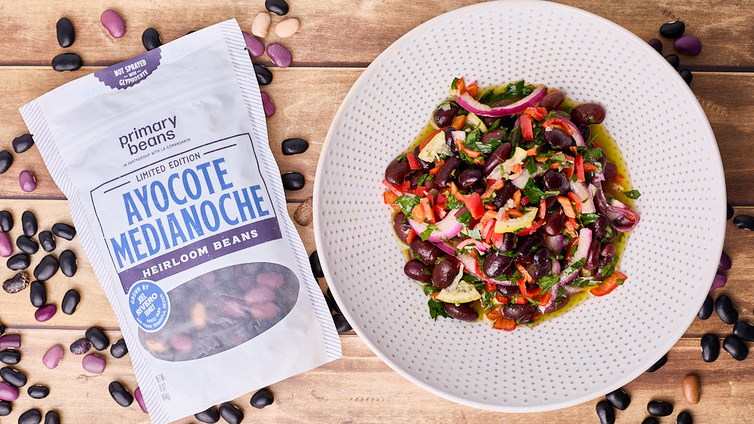 A bag of ayocote medianoche heirloom beans next to a colorful bowl of beans with vegetables