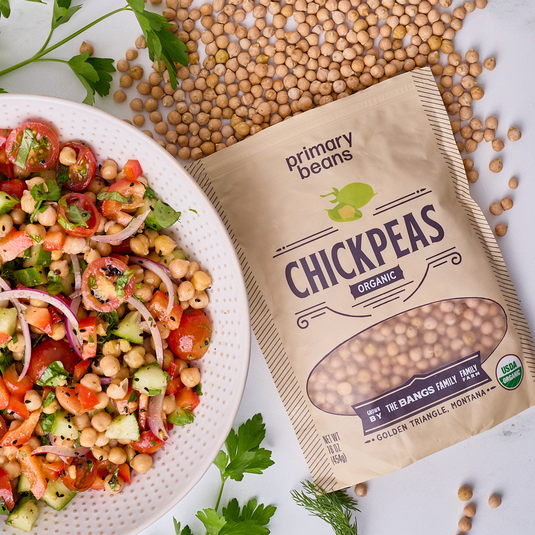 A bag of Primary Beans Organic Chickpeas lies on a white surface beside a white plate of fresh chickpea salad, with scattered whole chickpeas around the package.