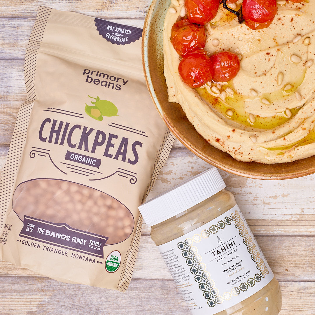 A bag of Primary Beans Organic Chickpeas next to a jar of Tahini, with a bowl of hummus topped with roasted tomatoes, arranged on a light wood background.