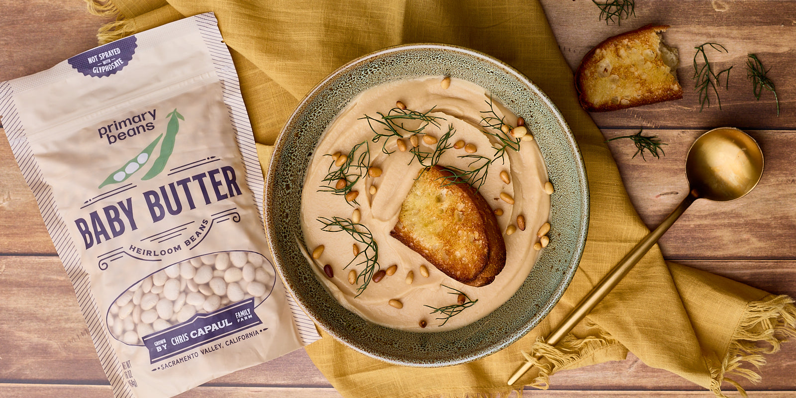 bowl of creamy white soup with a piece of grilled bread and herbs next to a bag of baby butter beans