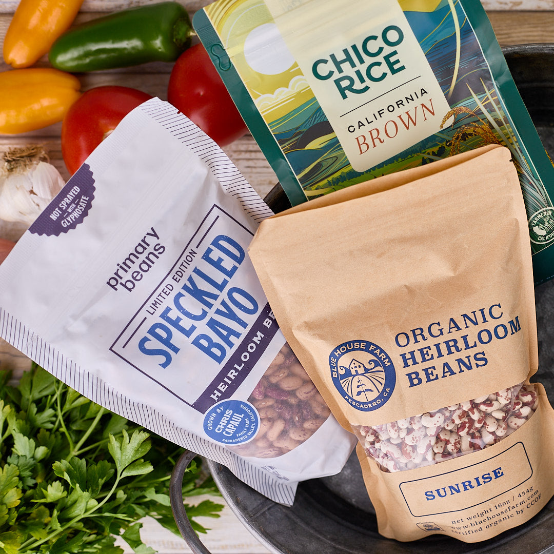 Three packages of beans on a wooden surface with tomatoes and herbs.