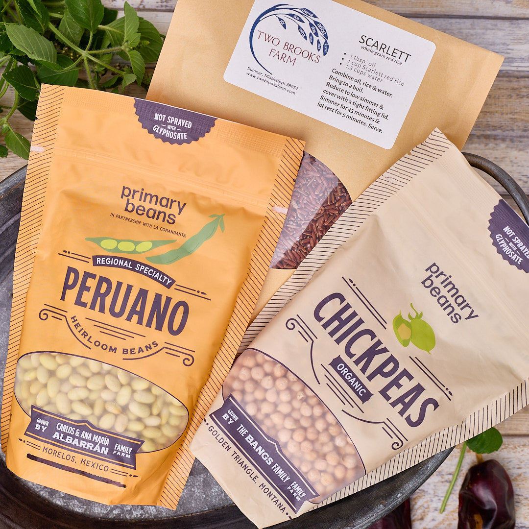 Two bags of Primary Beans organic legumes on a rustic wooden surface with a metal tray, garnished with green herbs. One bag is a golden-yellow color labeled 'Peruano', and the other is a light beige color labeled 'Chickpeas' (garbanzo beans).