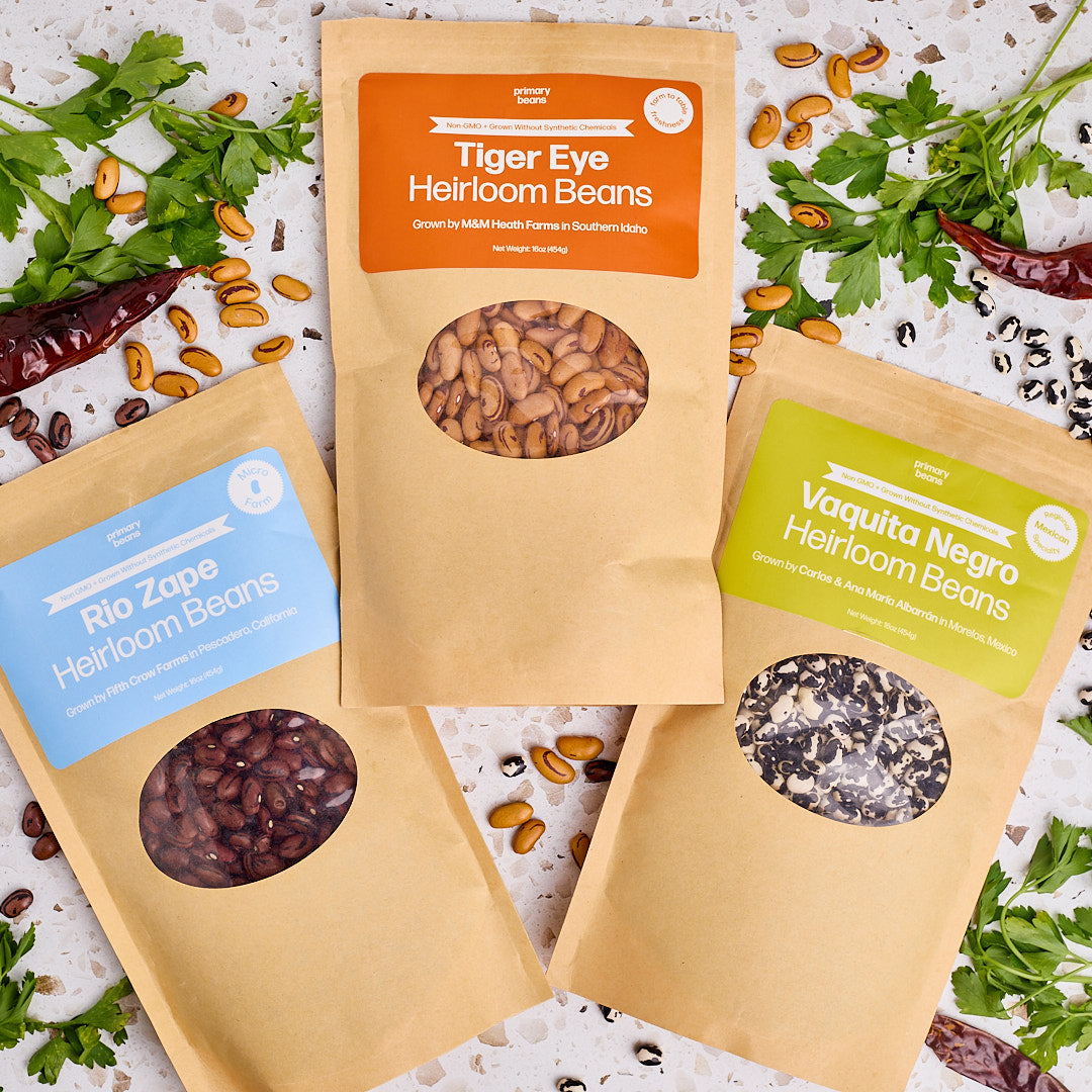 3 packages of heirloom beans on counter with herbs and chili peppers around.