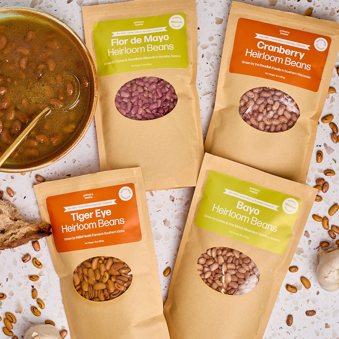 Four packages of heirloom beans with visible labels on a textured surface with beans and soup.