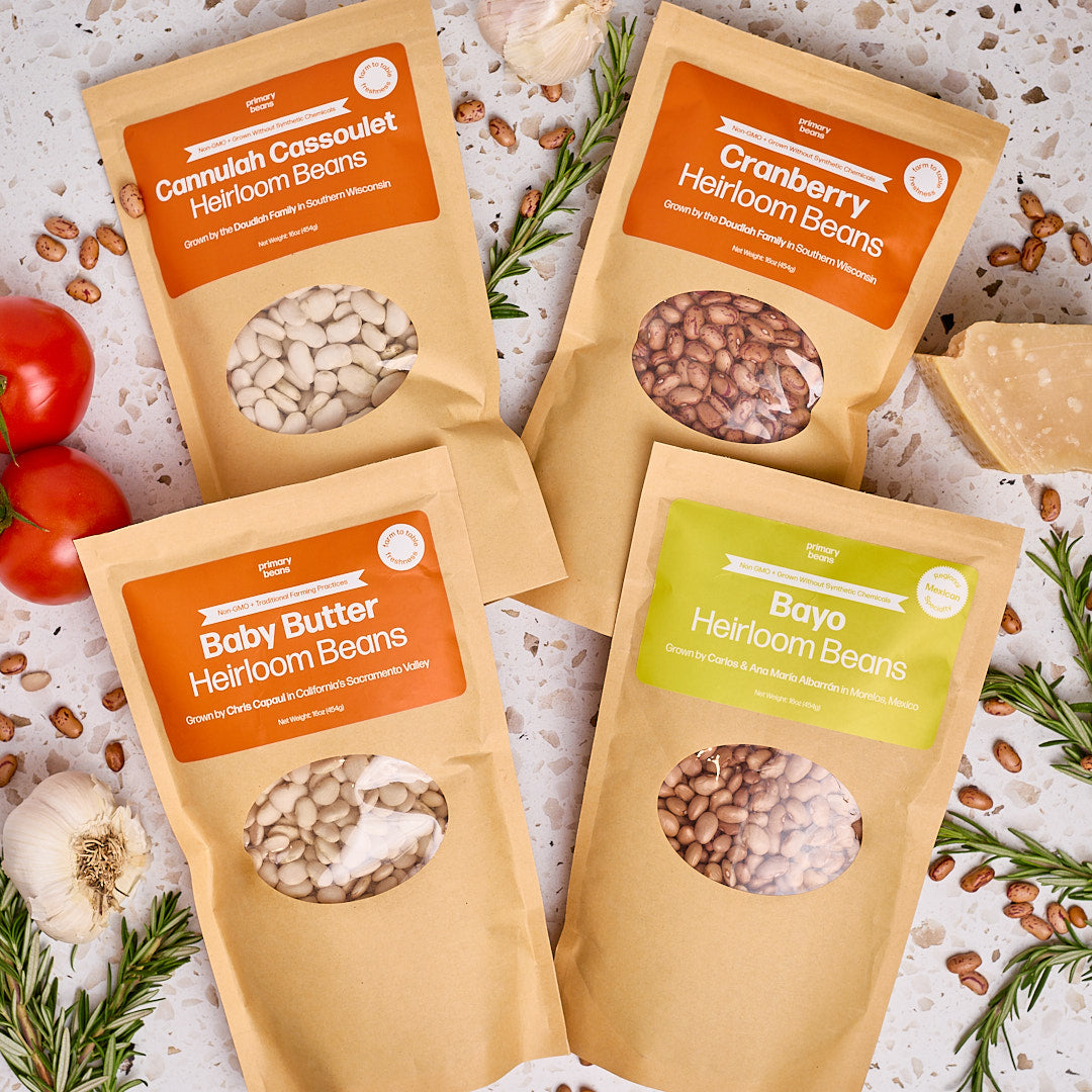 Four packages of different types of heirloom beans on a textured surface with herbs and tomatoes.