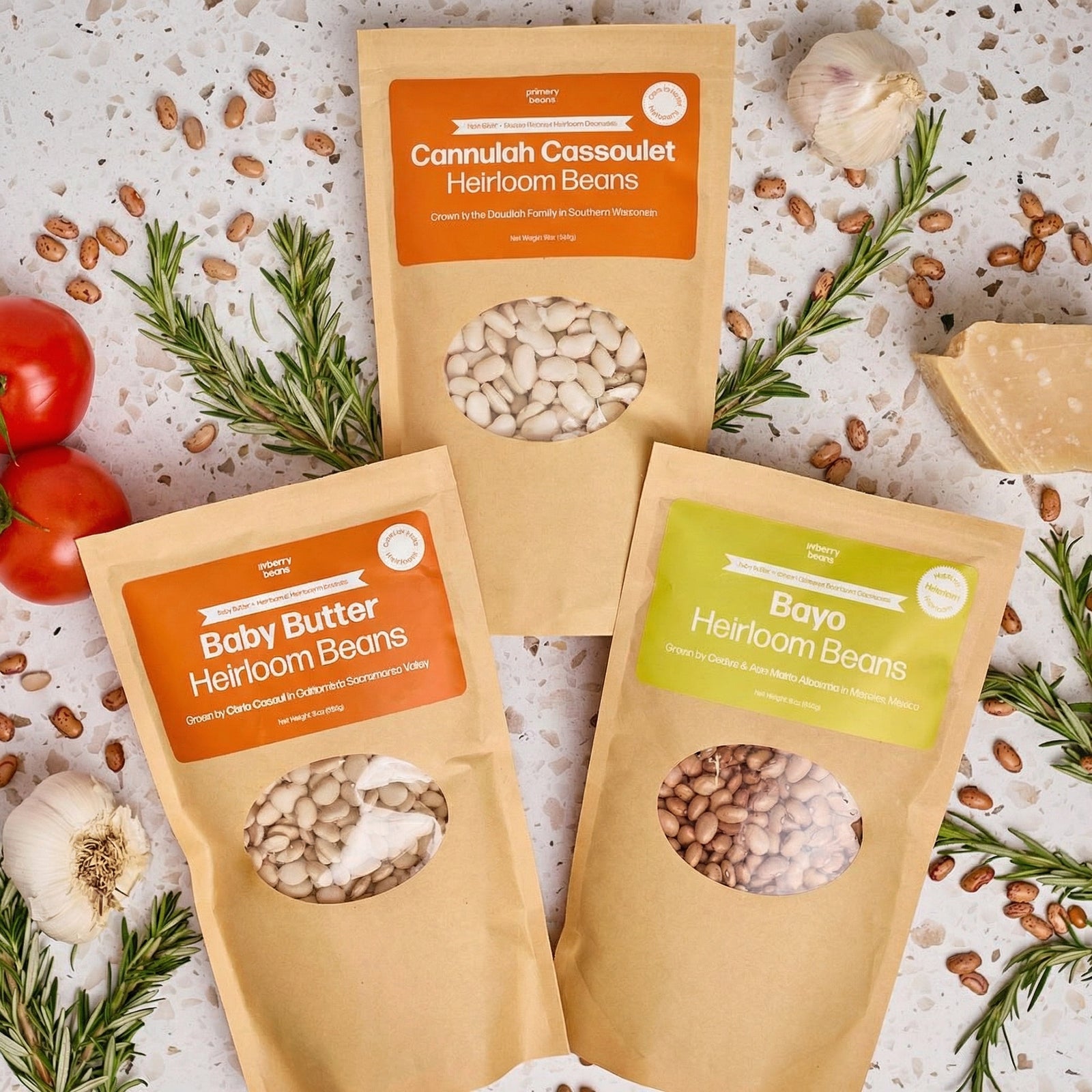 Three packages of heirloom beans with visible labels on a textured surface with herbs and tomatoes.