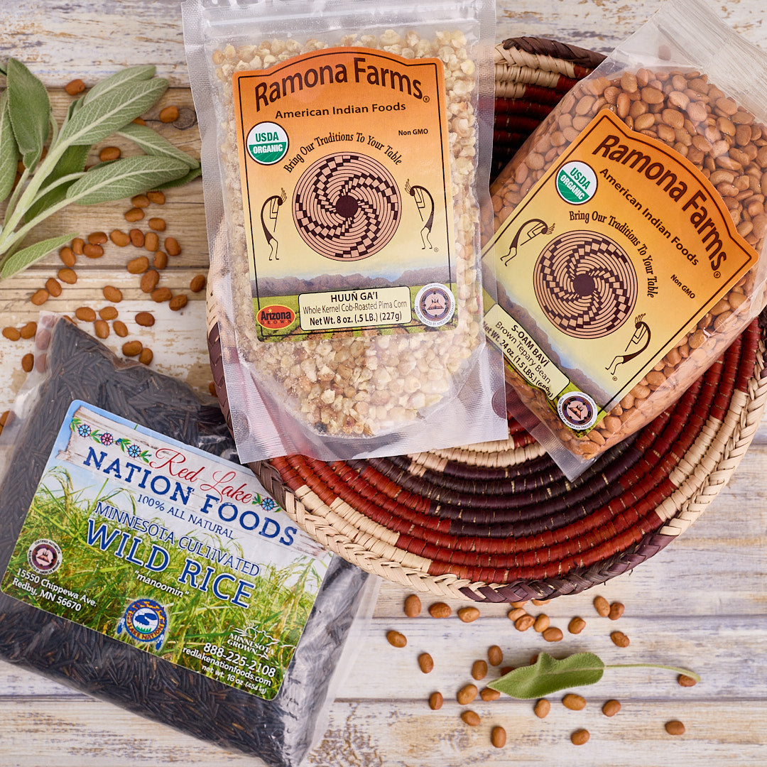 Packages of Ramona Farms tepary beans and Red Lake Nation Foods indigenous wild rice on a wooden surface with beans and leaves.