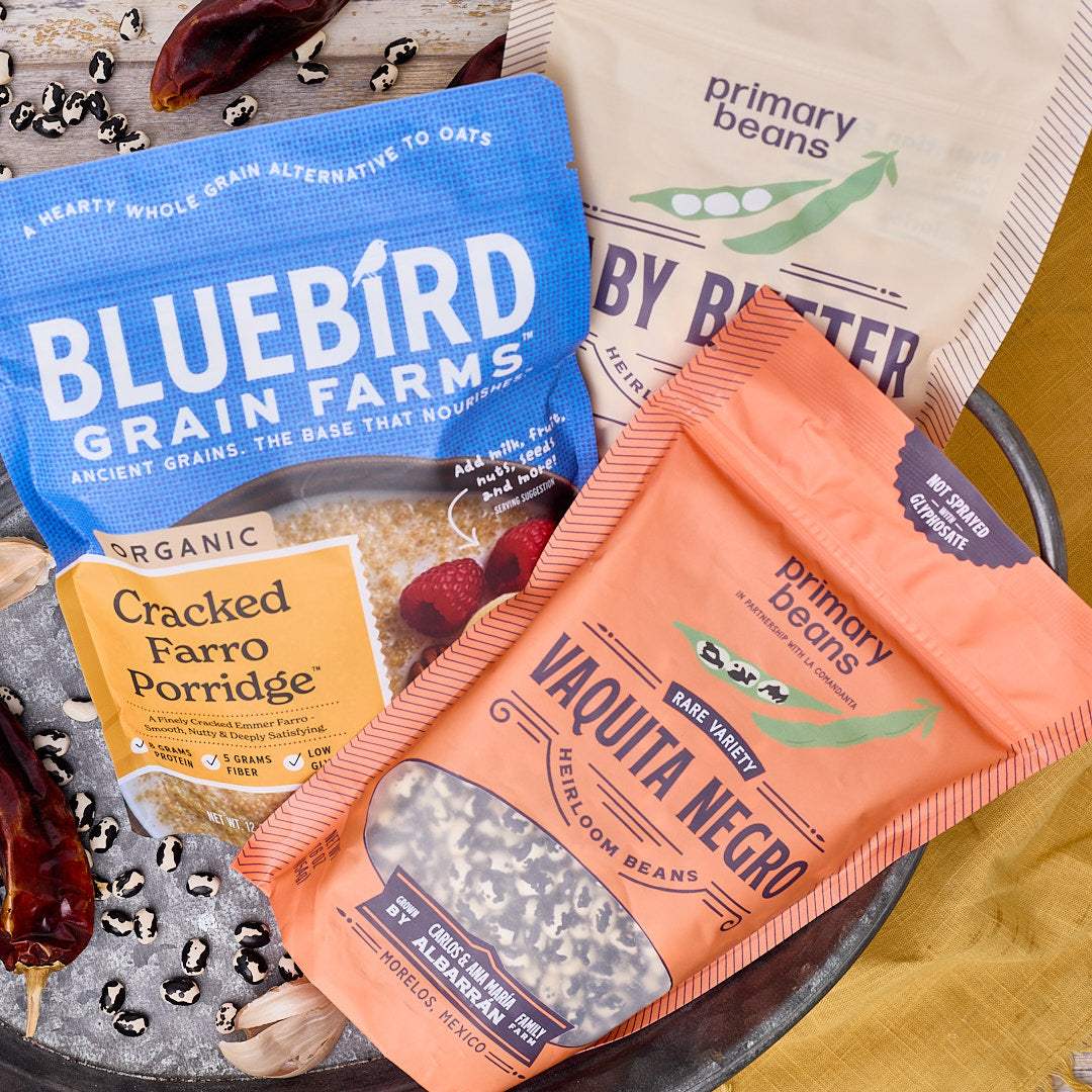 A styled flat-lay of several grain and bean product bags arranged on a dark tray with decorative dried chilies and garlic. Visible brands include Bluebird Grain Farms and primary beans.