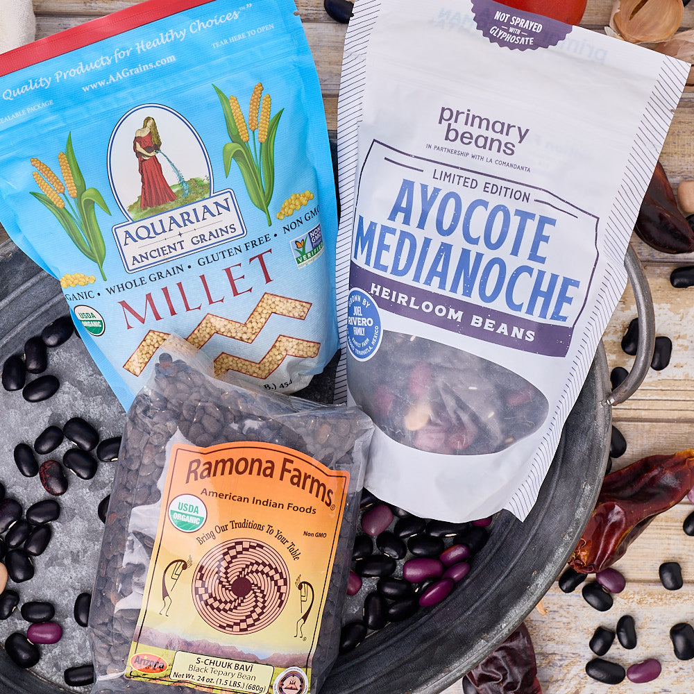 two bags of heirloom beans and one bag of millet in a tin pale sitting on a wood surface decoratively scattered with colorful beans and chilis