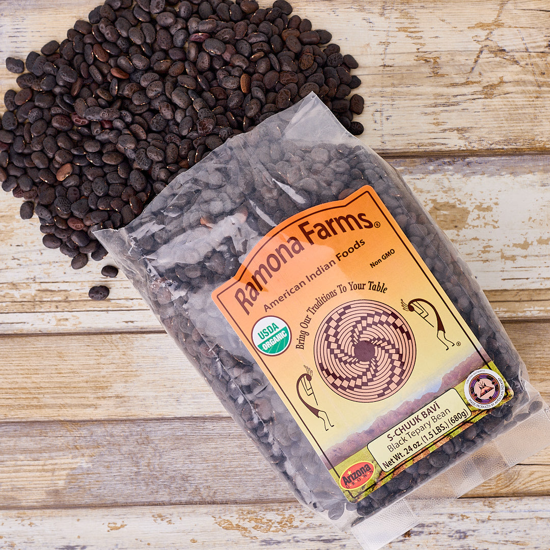 A clear plastic bag of Ramona Farms S-Chunk Savi dried tomatoes, with loose dark red tomatoes spilling out onto a light wood surface. The bag features an orange and yellow label with a USDA Organic seal.