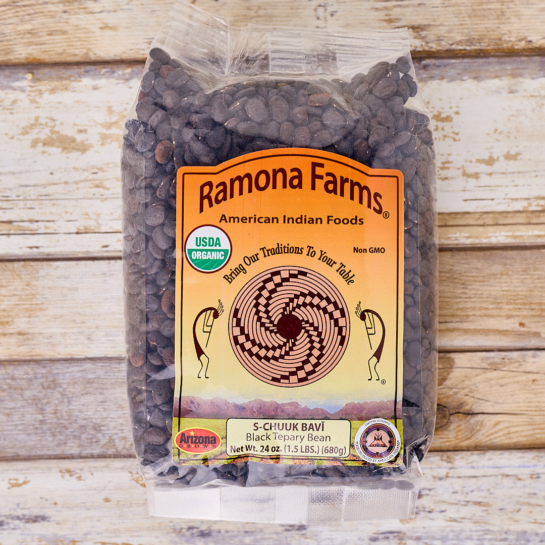 A 24 oz bag of Ramona Farms S-CI-UUK Bavi Black Tsoory Bean, shown on a rustic whitewashed wooden background. The clear plastic bag contains dark brown dried beans and features a prominent orange and yellow label with the Ramona Farms logo.