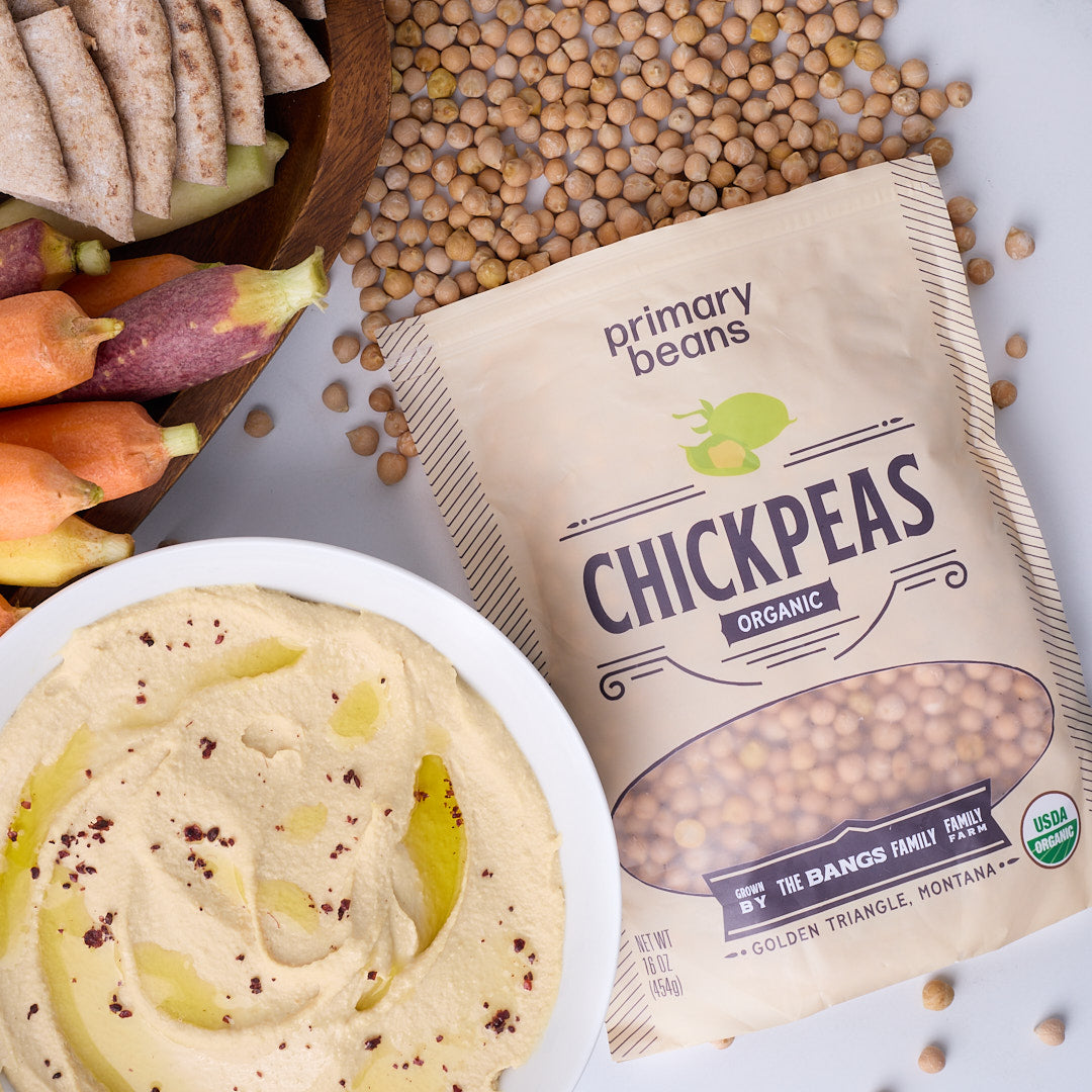 A bag of Primary Beans Chickpeas Organic next to a bowl of hummus with vegetables, set on a white surface with scattered chickpeas.