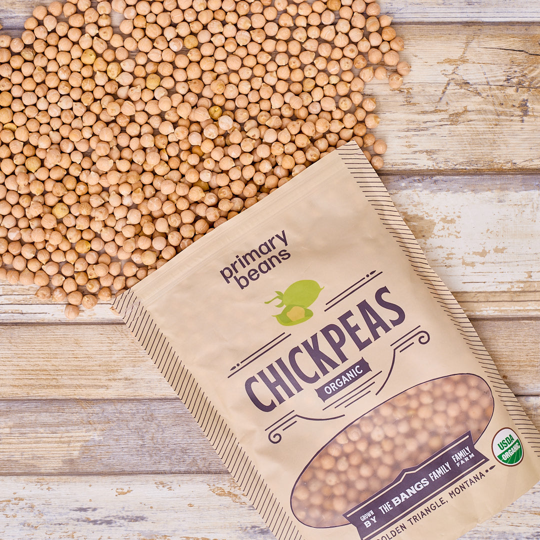 Bag of organic chickpeas from Primary Beans on a wooden surface with chickpeas scattered around.