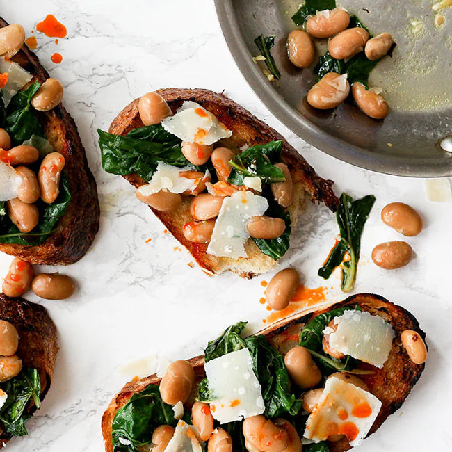 Toasted bread topped with heirloom beans, sautéed greens, shaved cheese, and a drizzle of chili oil on a marble surface.